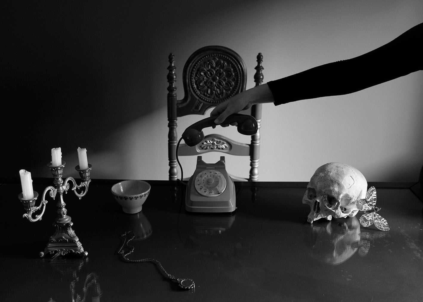 A black and white poster featuring a still life arrangement with a skull, an antique chair, a telephone, candle holders, and a bowl on a table.