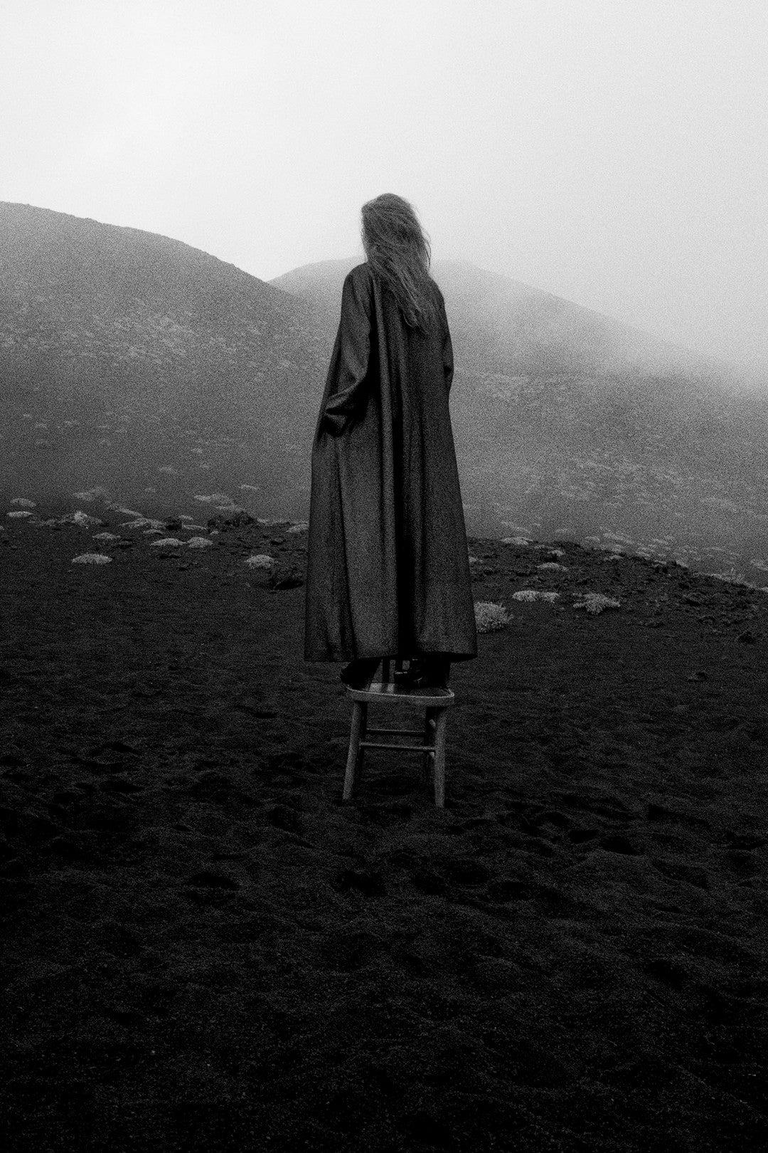 Person in a long coat standing on a stool in a foggy, mountainous landscape