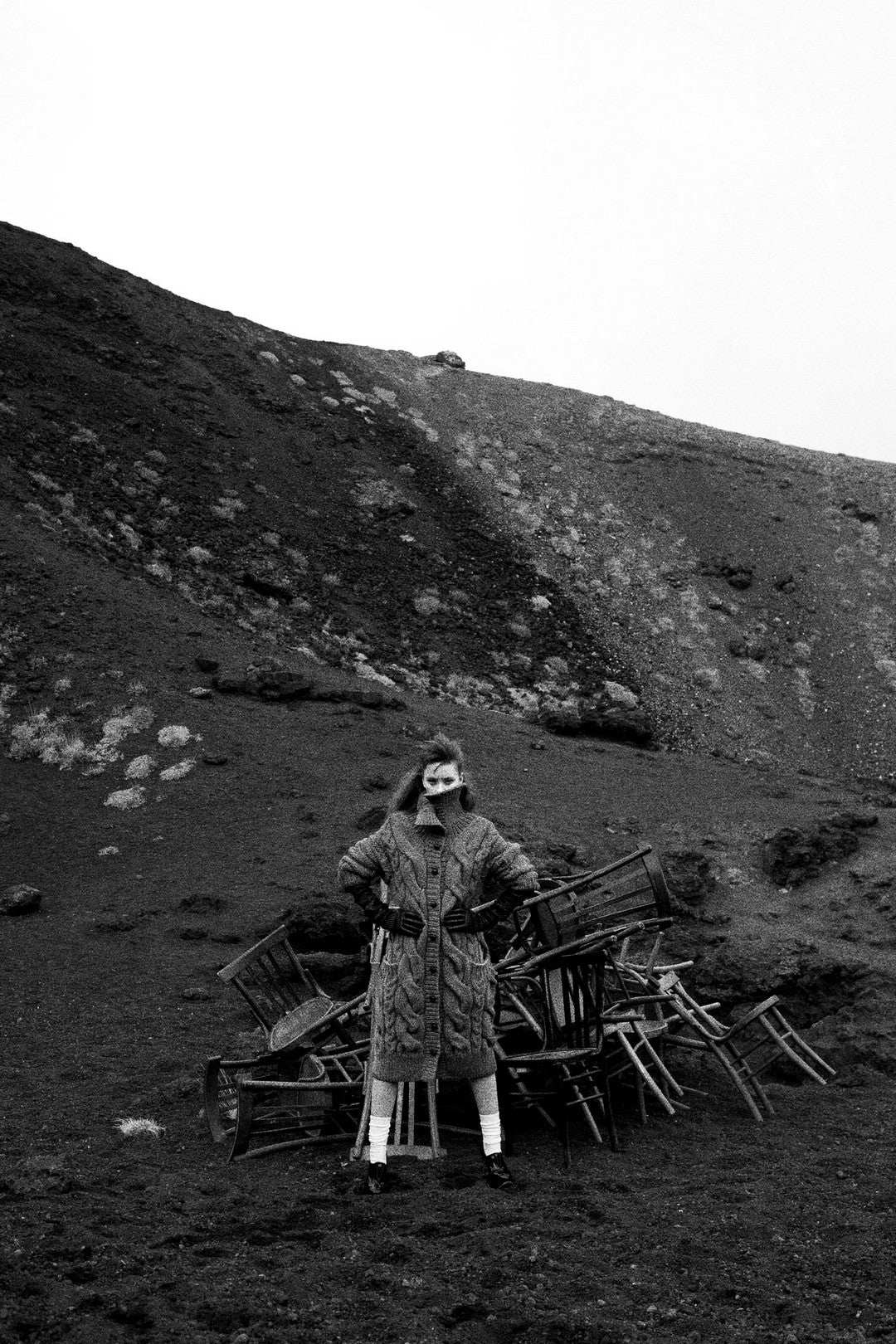Person standing in a rugged landscape with chairs, wearing a coat and hat.