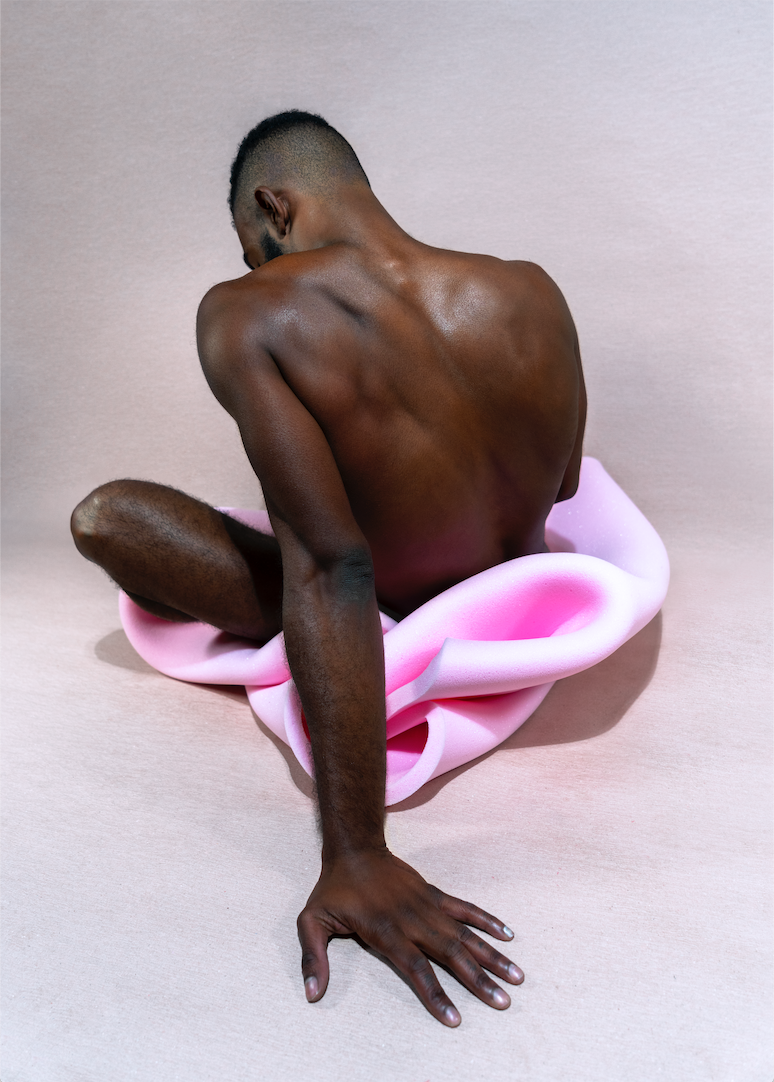 This image presents "Florian Hetz #2," a fine art photograph by Florian Hetz, capturing the fragmented and subjective experience of encountering another man's body. The photograph features a close-up view of a male figure seated, with a focus on the play of light and shadows across the figure's back and the soft, pink-colored object he interacts with.