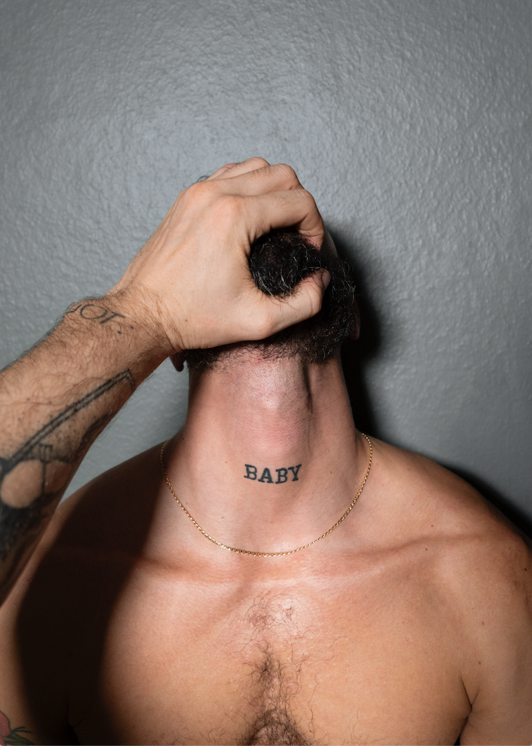A black and white image depicting a close-up of a person's face and chest with a tattoo, wearing a gold necklace.