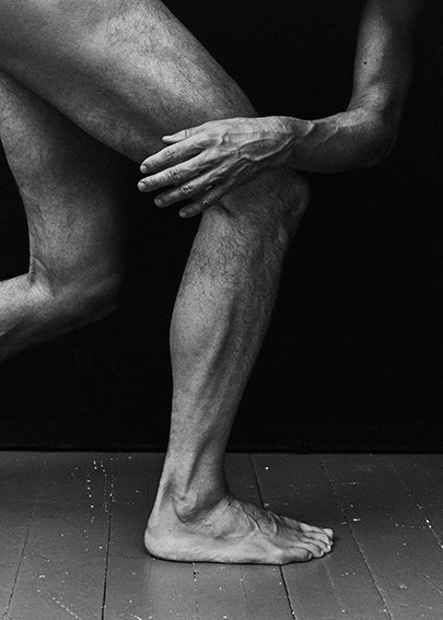 Black and white photograph of a human leg resting on a foot, set against a dark background, highlighting textures and shadows.