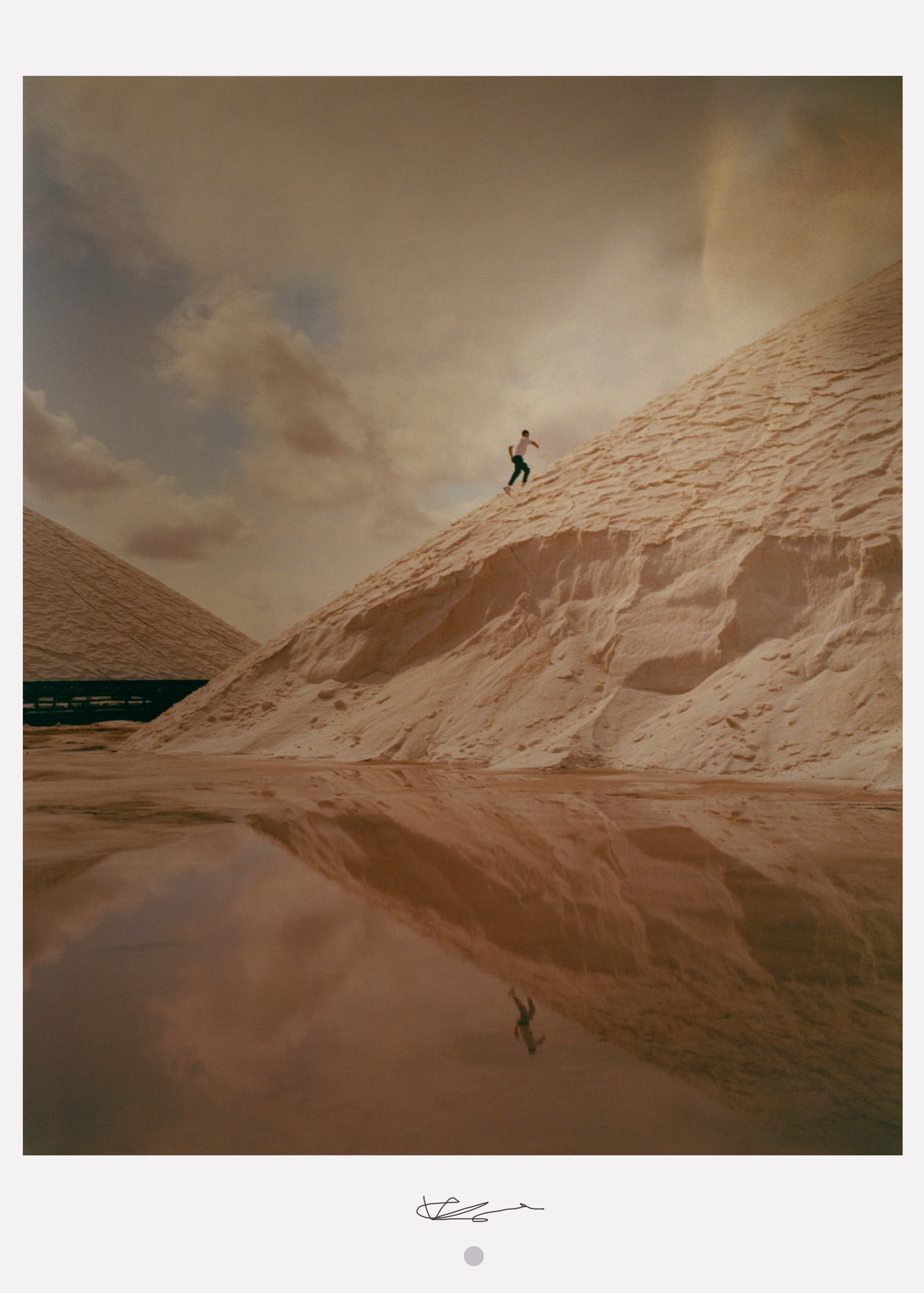 A framed art print featuring a person standing on a sand dune with a body of water and a cloudy sky in the background.